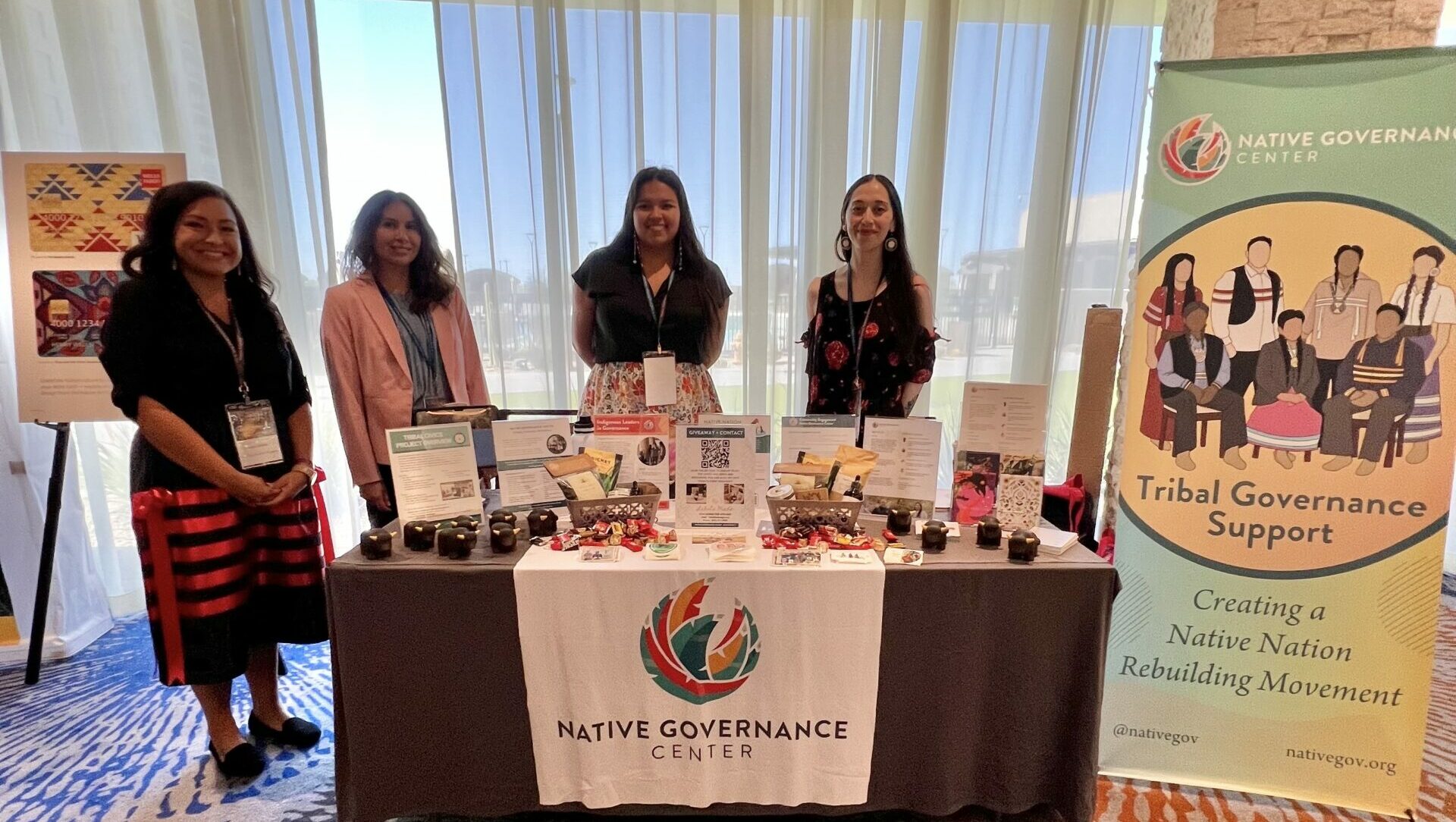 4 women stand behind a colorful table for Native Governance Center at last year's WEWIN conference.