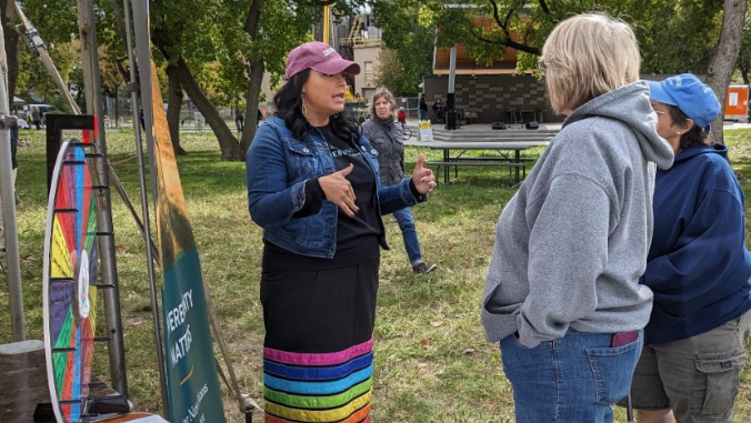 NGC Staff tabling outside on grass at previous fair event in colorful ribbon skirt talking to two fair attendees.